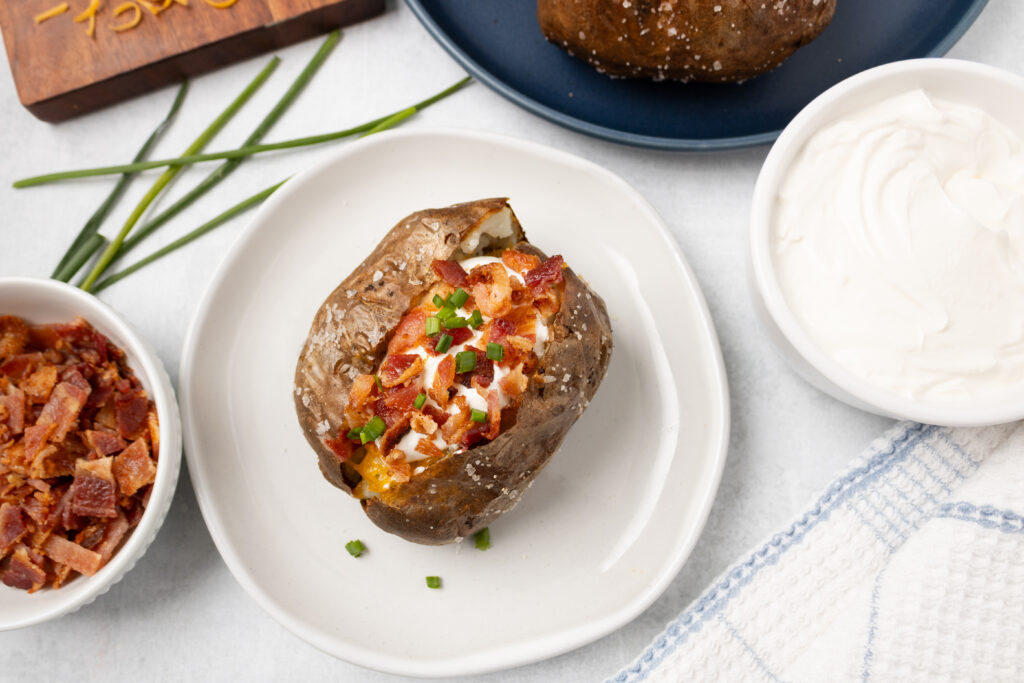 An overhead picture of the air fried baked potato with bacon, cheddar cheese, sour cream and chives.  There is a bowl of sour cream and a bowl of bacon bits .