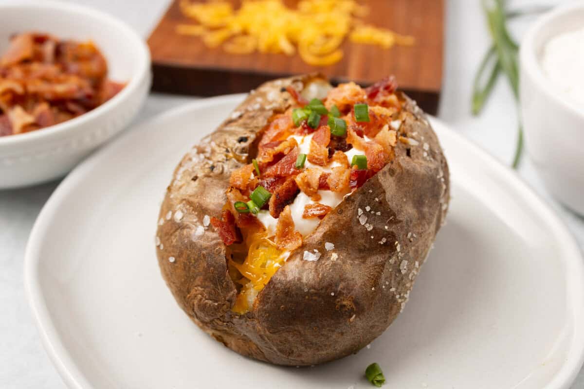 Front potato is split open wth bacon, cheddar cheese, and sour cream. Background has bacon.