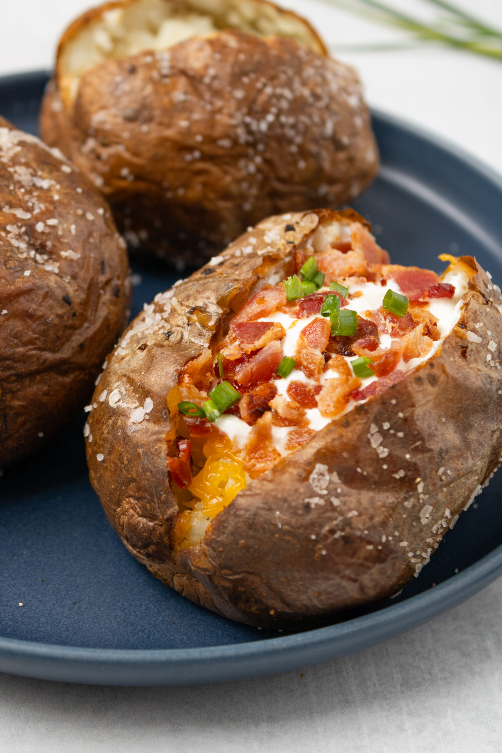 Three air fried baked potatoes coated with kosher salt on the outside,  Front potato is split open wth bacon, cheddar cheese, and sour cream