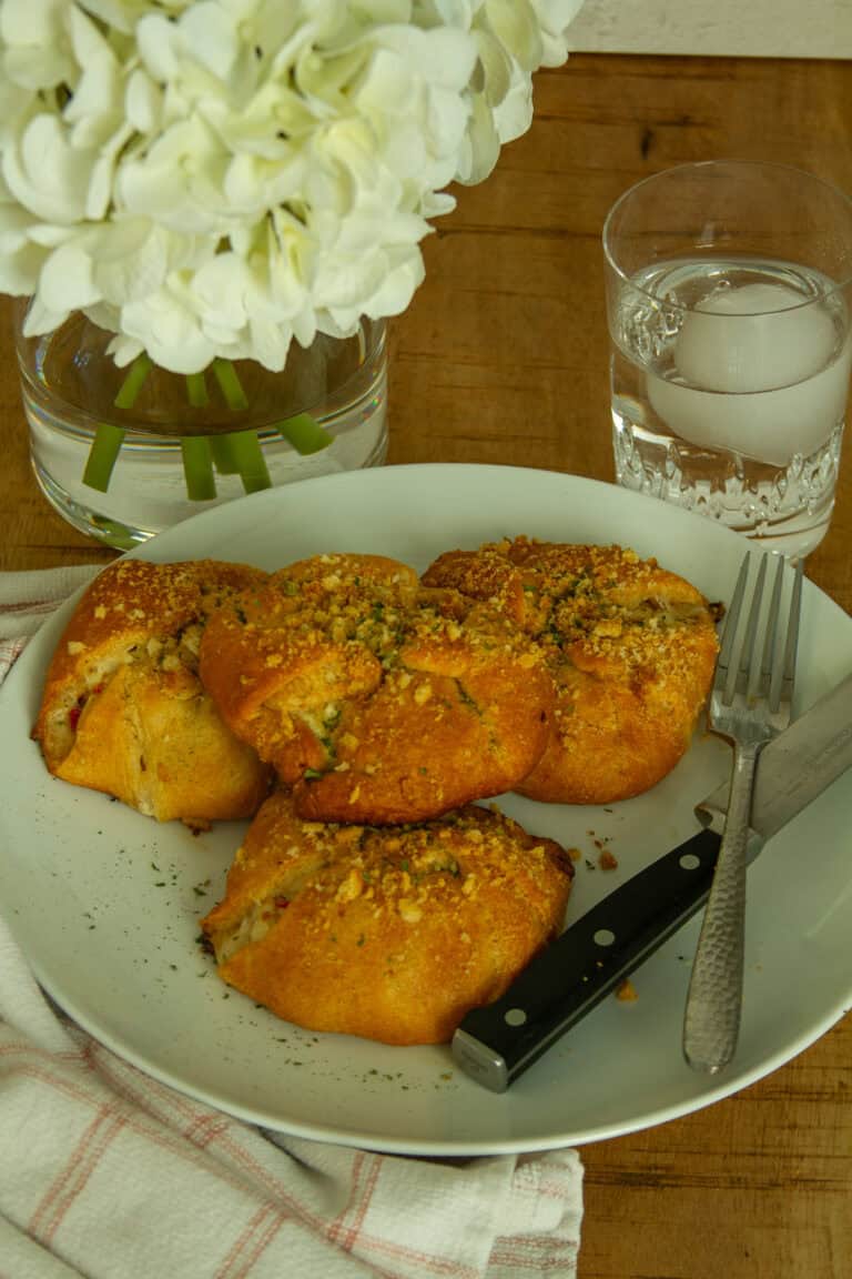 Four golden Chicken Crescent Rolls on a white plate with a fork, a glass of water, and white flowers in the background.