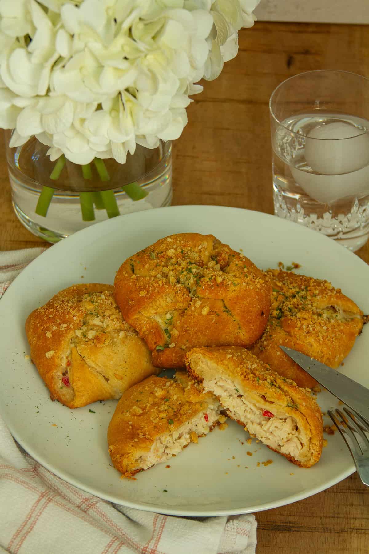 Picture of 4 chicen crescent rolls with a knife and fork glass of water and a beautifulfloral arrangment. One of the chicken crescent rolls is cut in half showing the layers of chicken and vegtables.
