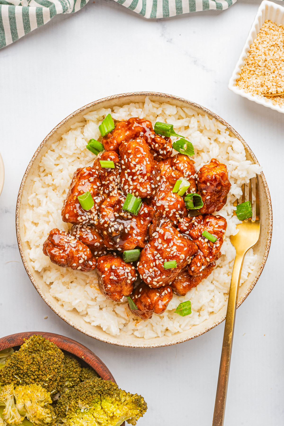 This is an over head shot over the dish.  The plate of food is bright vibrant colors of orange chicken covered in white sesame seeds and green onion,  Served on a bed of white rice accompanied by a gold fork.