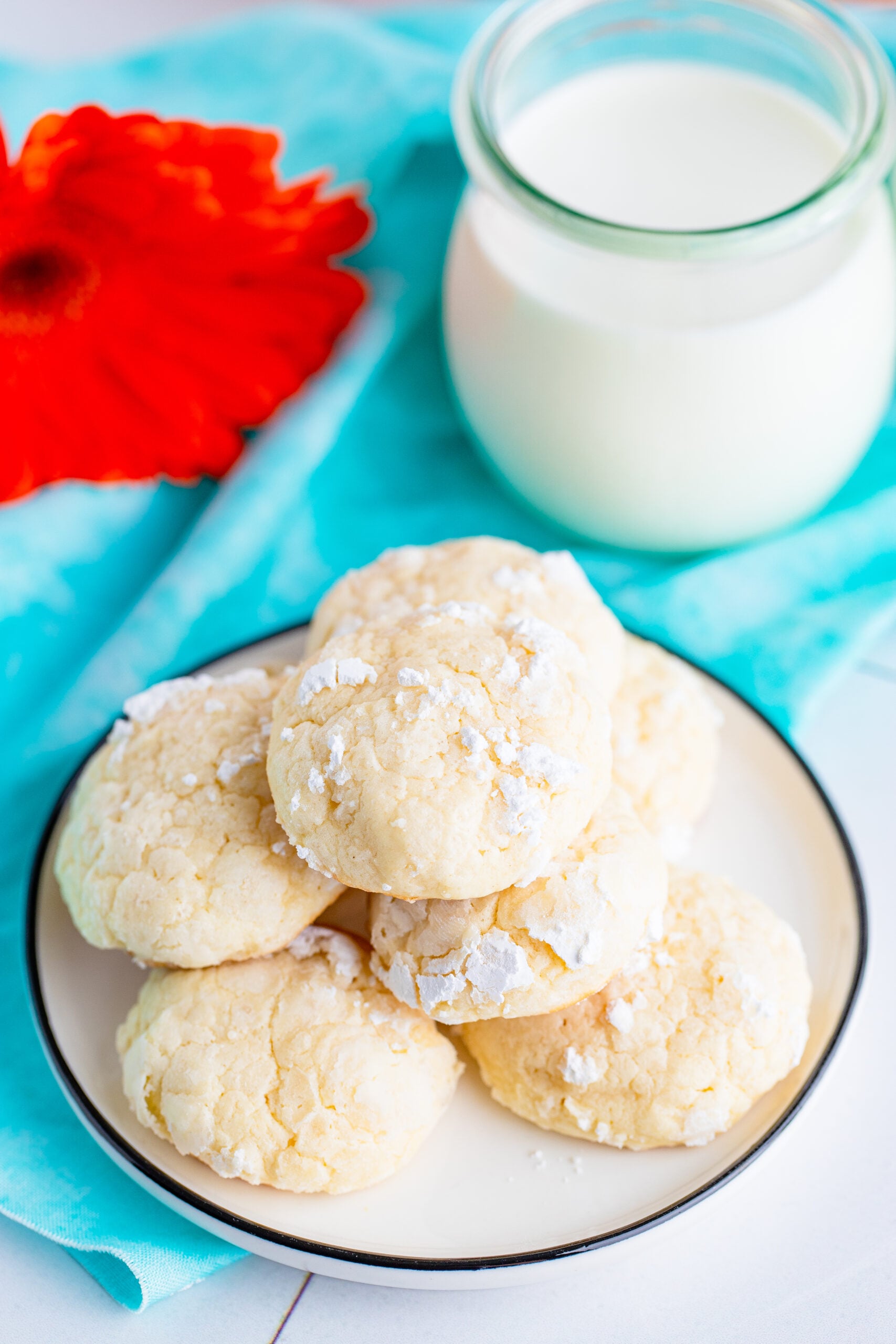 This is a picture of the cream cheese cookies stacked on top of each other on a plate resting of a blue napkin that offsets the white cookies and a glass of milk in the background.