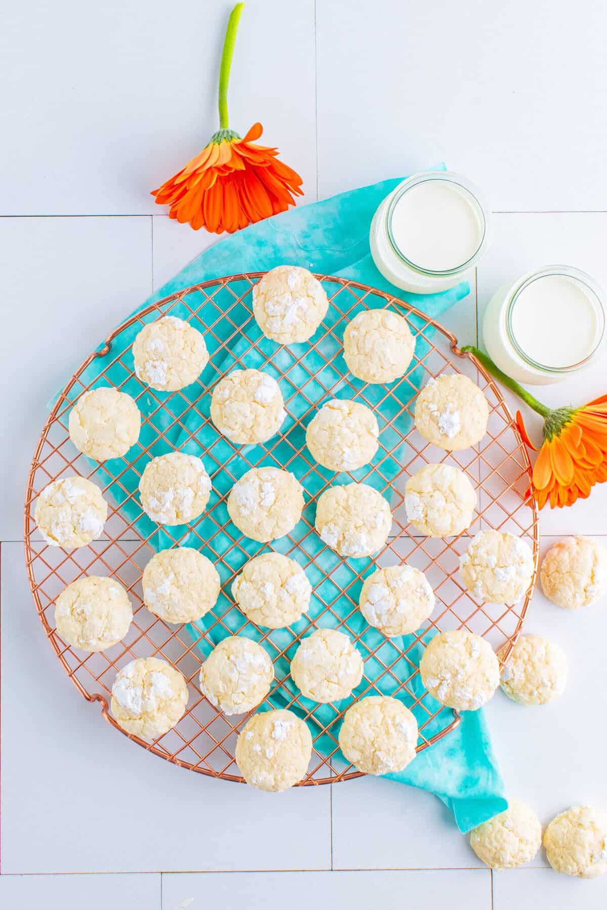 A picture of cream cheese cookies on a metal tray with a blue scarf underneath. There are two glasses of mike and two orange flowers.