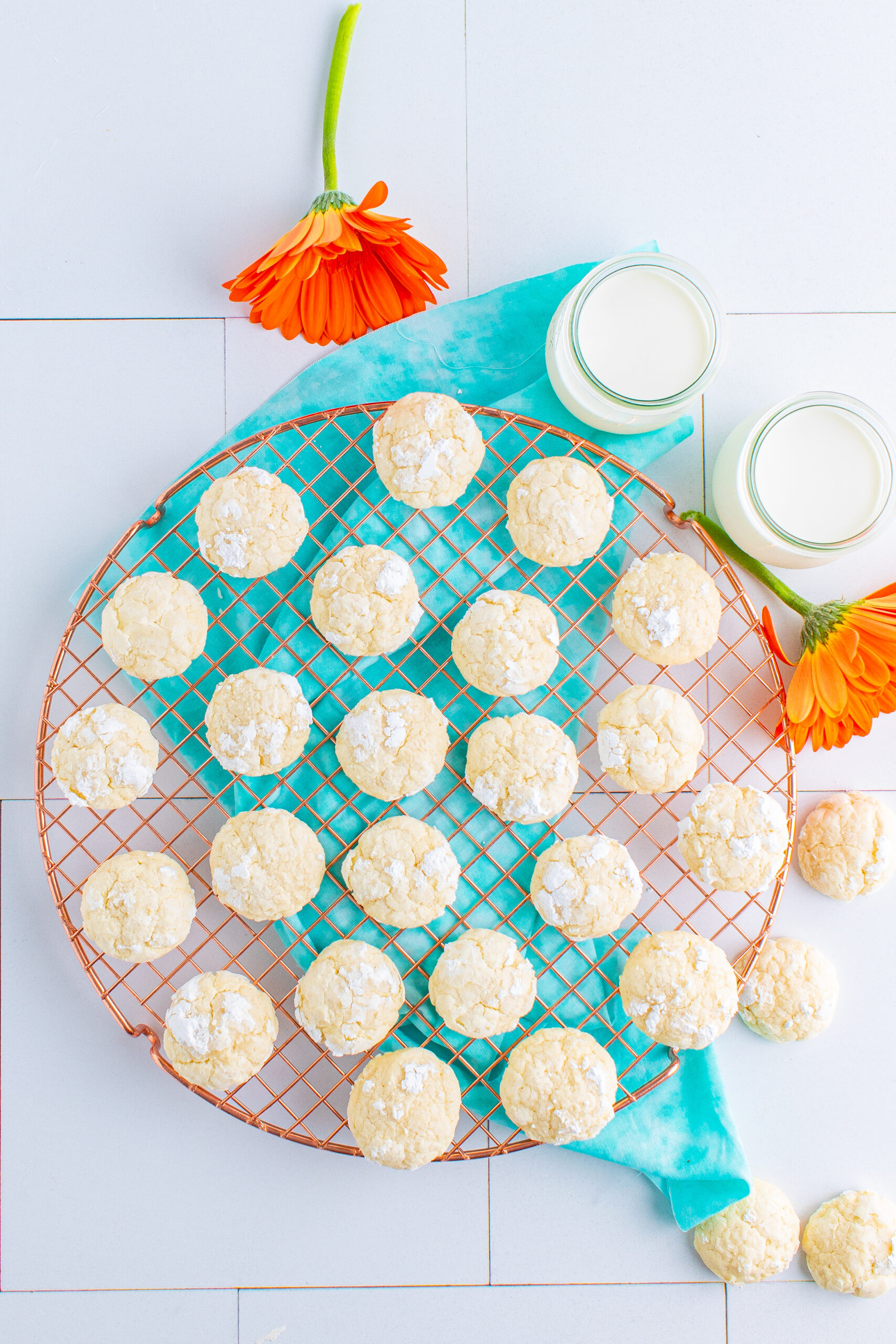 A picture of cream cheese cookies on a metal tray with a blue scarf underneath.  There are two glasses of mike and two orange flowers.