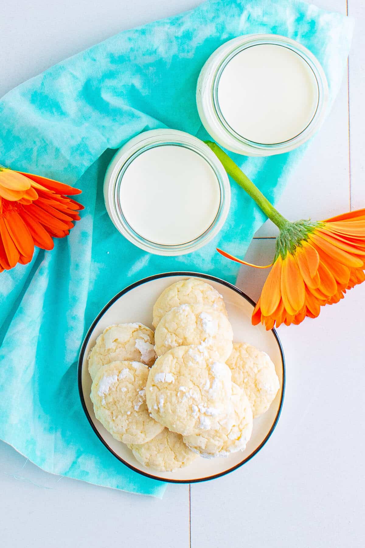 This is a picture of the cream cheese cookies stacked on top of each other on a plate resting of a blue napkin that offsets the white cookies and a glass of milk in the background. This picture has swo orange flowers in it.