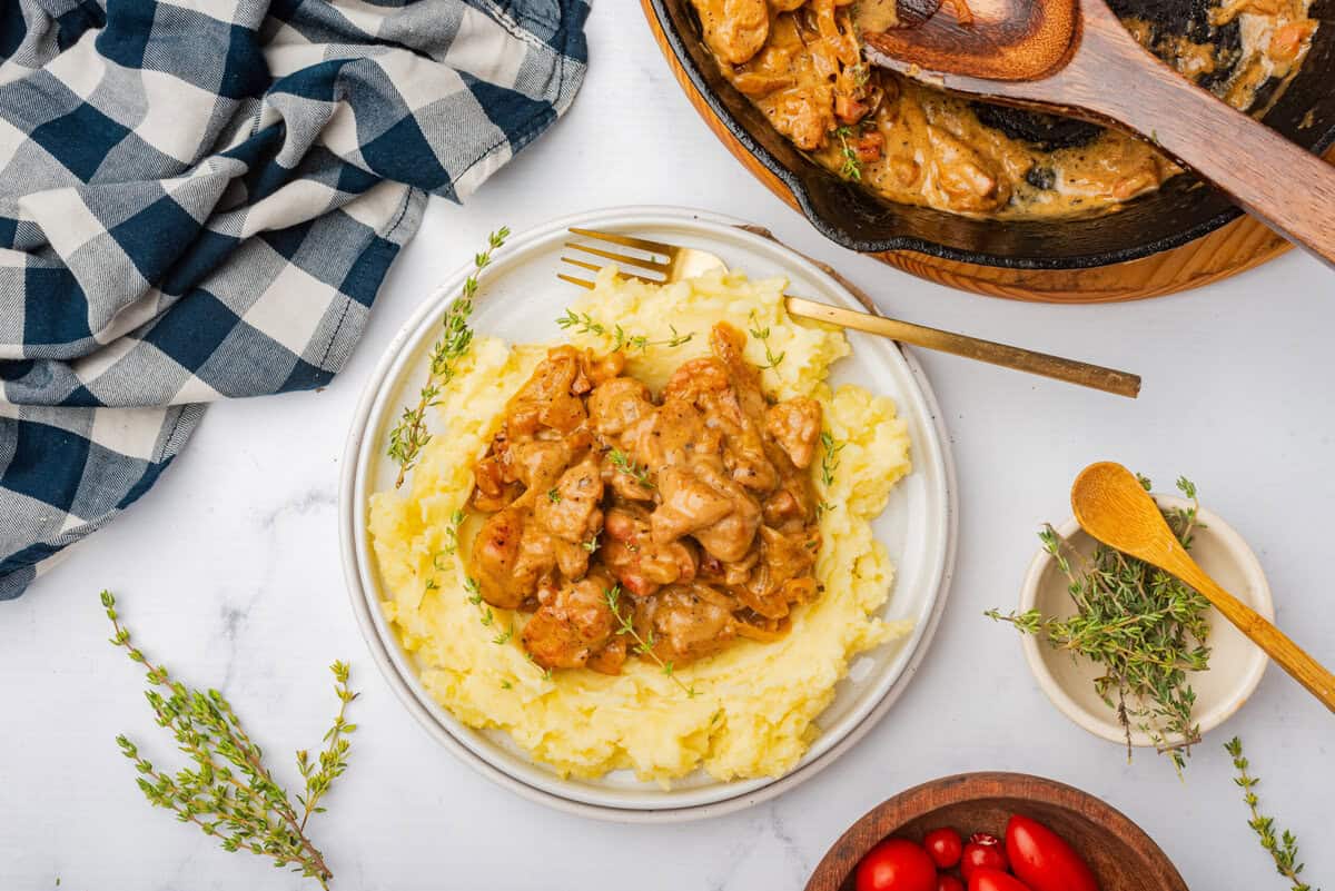 This is an overhead picture of the sweetbreads in the sauce created in the recipe served on a bed of mashed potatoes surrounded by the pan used to create the recipe thyme sprigs and a bowl of tomatoes.