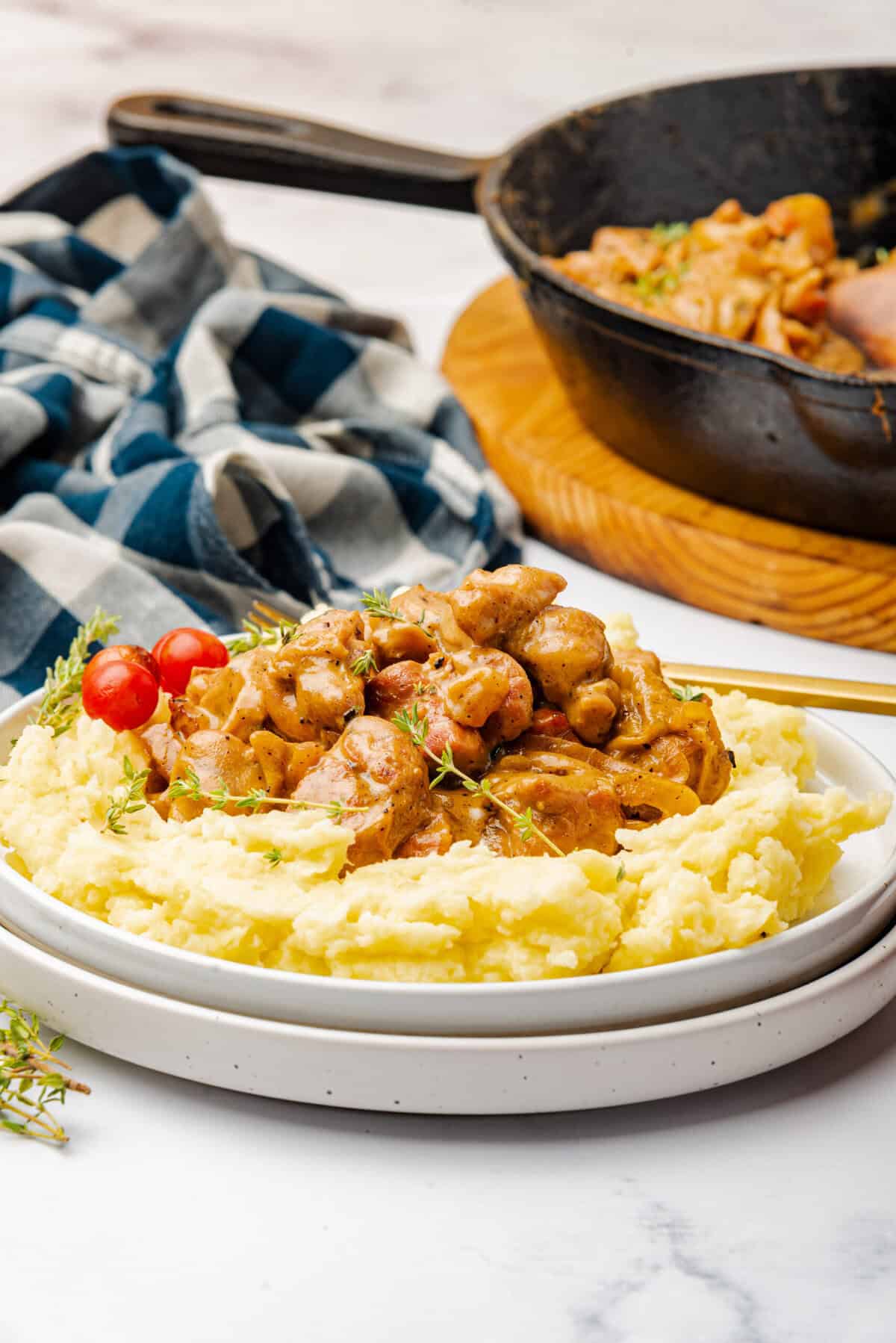 A plate of sweetbreads that are cooked with the sauce included in the recipe. Covered with thyme sprigs and tomoatoes served on a bed of mashed potatoes. This picture also included the pan used to create the recipe and a blue and white checkered napkin.
