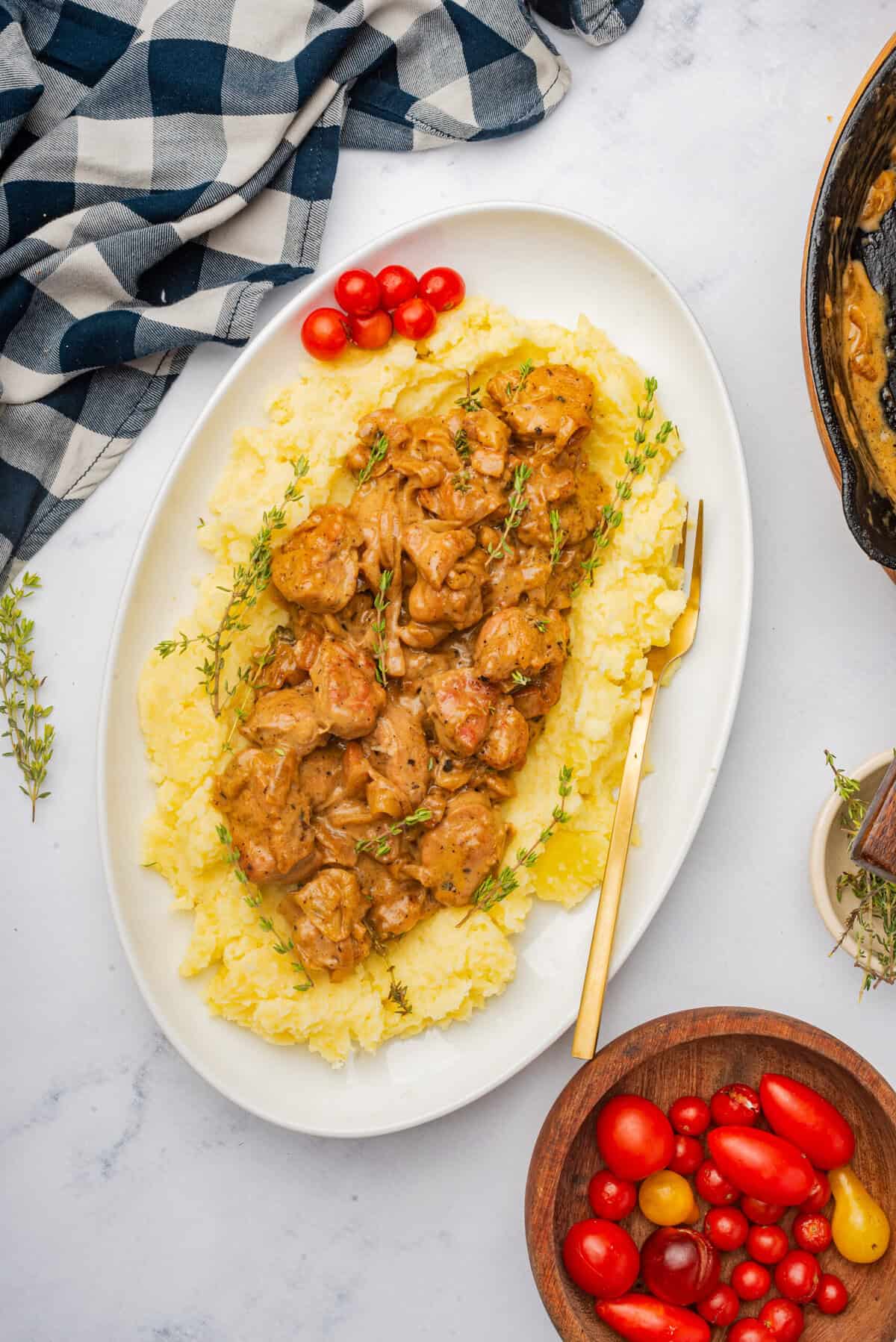 This is an overhead picture of the sweetbreads in the sauce created in the recipe served on a bed of mashed potatoes surrounded by the pan used to create the recipe thyme sprigs and a bowl of tomatoes. This is on an oval plate.