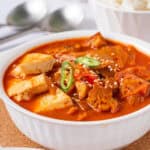 A bowl of spicy red kimchi jjigae with pork belly and tofu served with a bowl of white rice