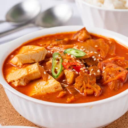 A bowl of spicy red kimchi jjigae with pork belly and tofu served with a bowl of white rice