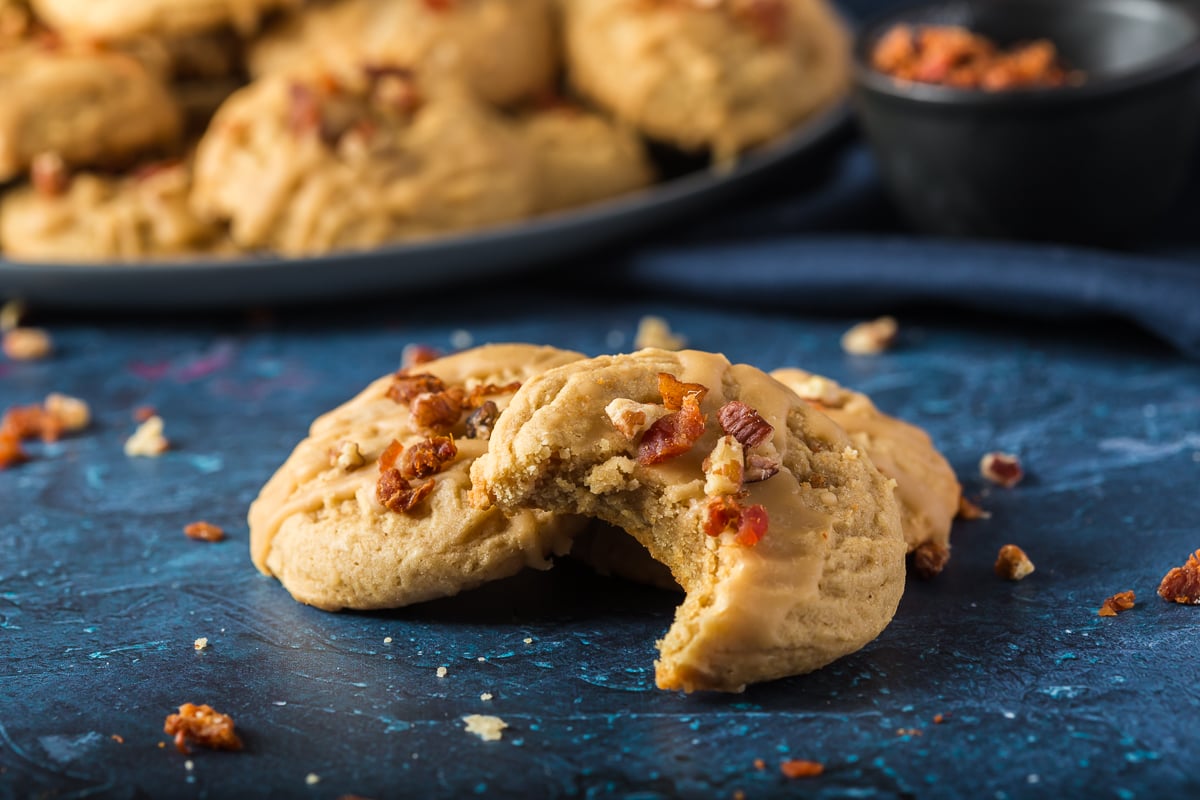 Final picture three cookies set up pyramid style with the first cookie having a bite out of it.  In the background there is a bown of bacon bits and a blate of maple bacon cookies.