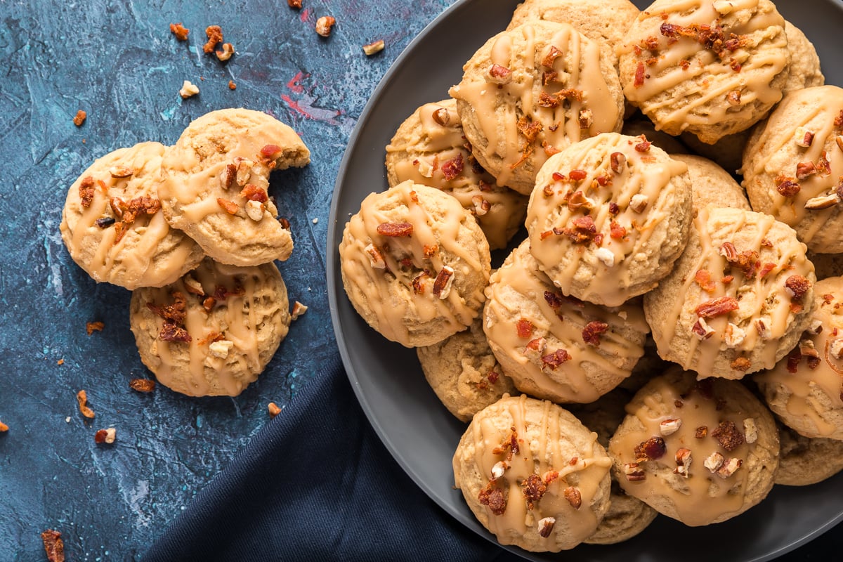 This is a picture of a group of maple bacon cookies.  There is a full plate of bacon maple cookies and three to the left of the plate one with a bite out of it.