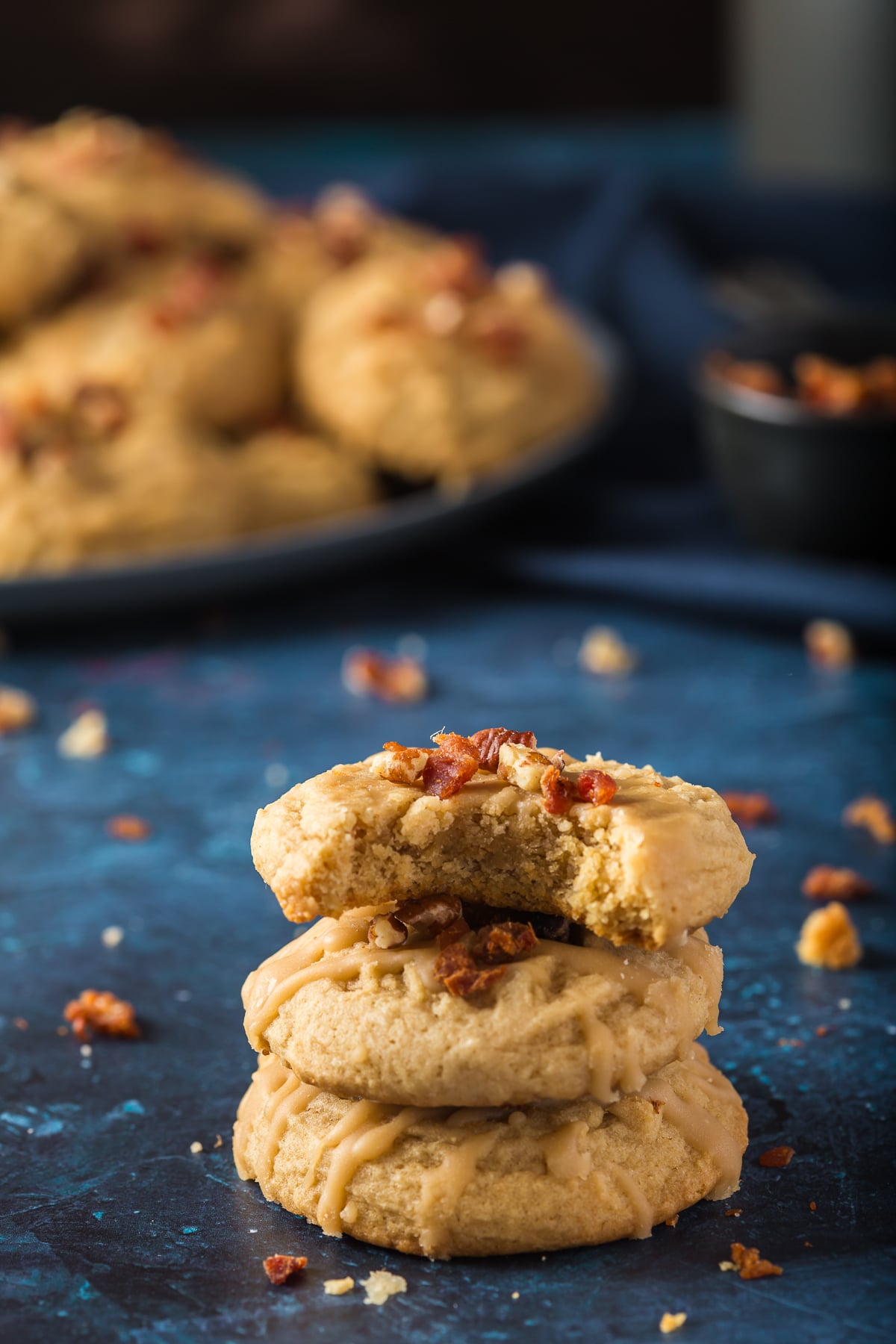 This is a picture of three maple bacon cookies stacked on top of each other.  Top cookie has a bite out of it.  In the background there is a plate of maple bacon cookies.