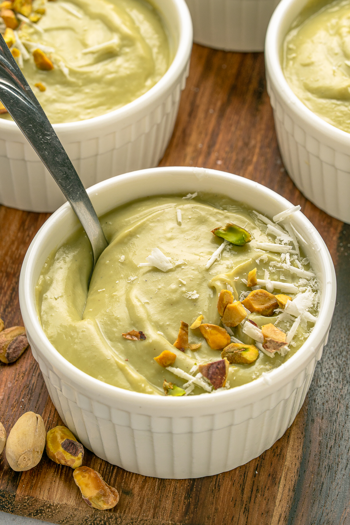 This is a picture of four bowls of pistachio pudding.  Decorated with crushed pistachios and white cheese in a white bowl.  They are on a wooden cake spinner.  This also has a spoon in the pistachio pudding ready to start the first bite.