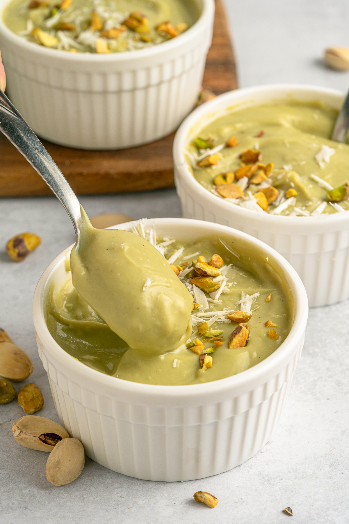 This is a picture of three bowls of pistachio pudding.  Decorated with crushed pistachios and white cheese in a white bowl.  They are on a wooden cake spinner. This has a picture of the spoon being pulled out of the bowl covered in pistachio pudding
