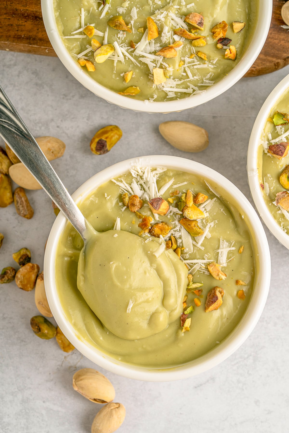 This is a picture of four bowls of pistachio pudding.  Decorated with crushed pistachios and white cheese in a white bowl.  They are on a wooden cake spinner.  This also has a spoon in the pistachio pudding pulling out the pudding.  This is an overhead shot looking at the dish in full glory.