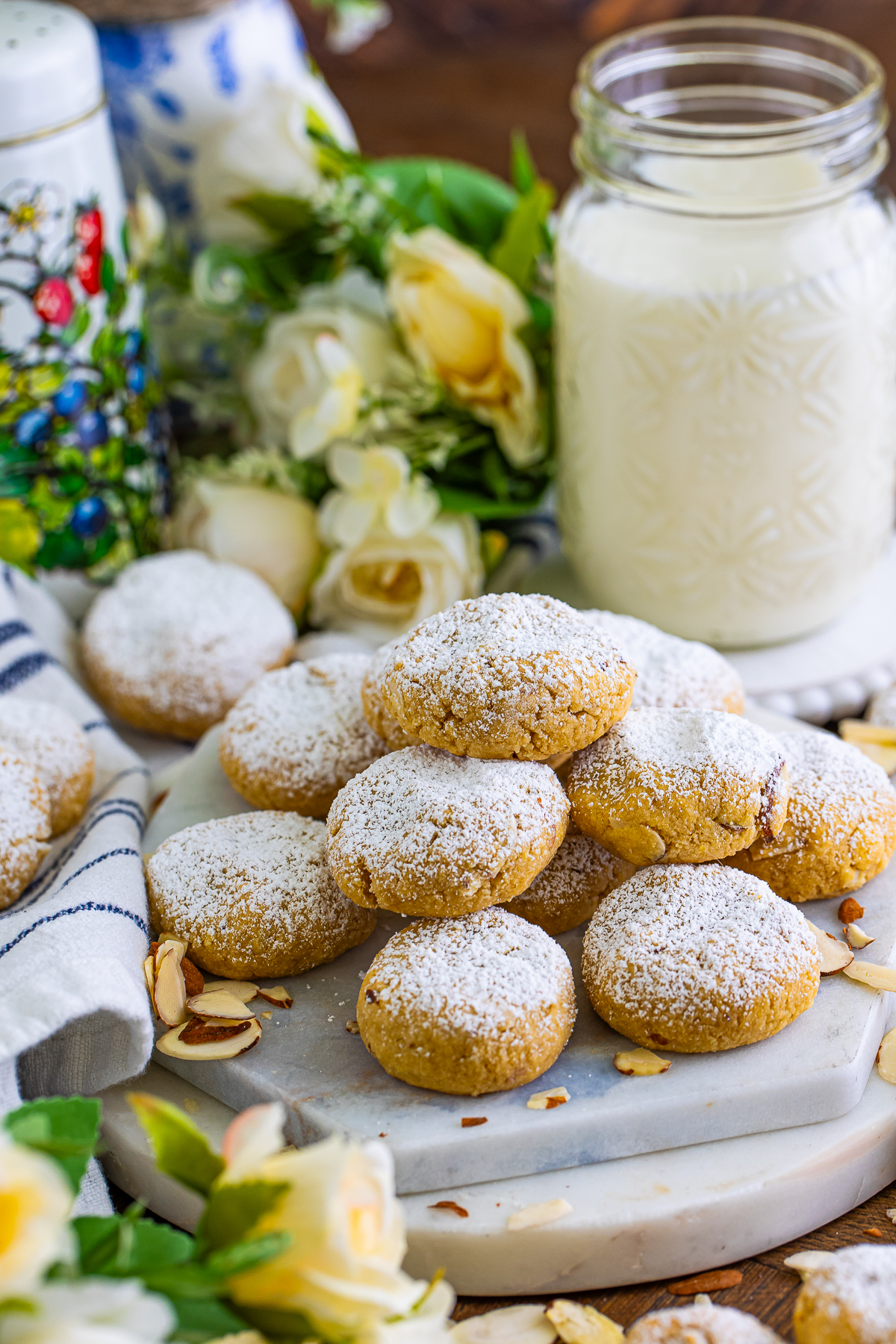 This is a picture of almond cookies covered with powdered sugar. They are stacked in a 4 dimensional pyramid with a glass of milk and yellow roses.