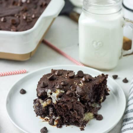 A gooey square of earthquake cake on a white plate with a jar of milk and chocolate chips scattered around.