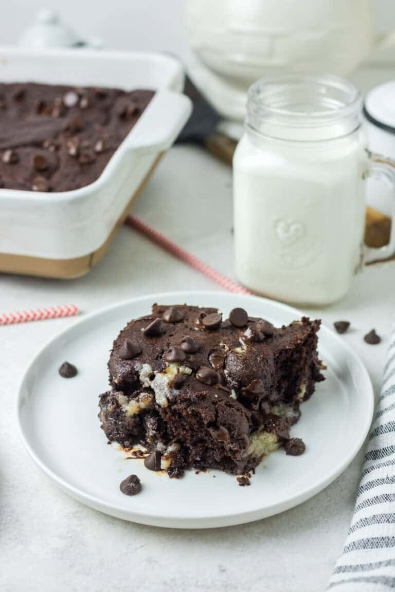 A gooey square of earthquake cake on a white plate with a jar of milk and chocolate chips scattered around.
