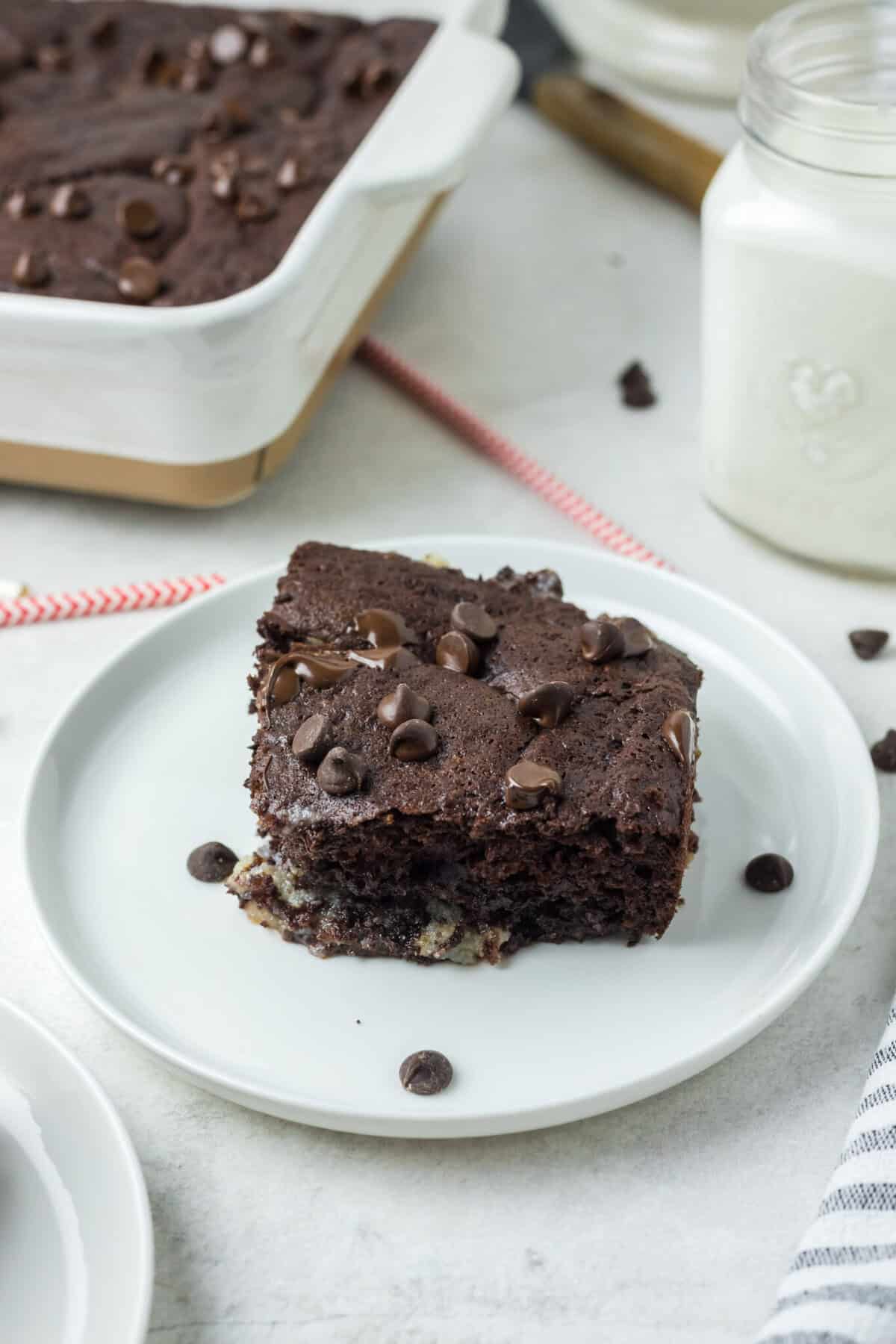 This is a pitcture of a piece of the chocolate earthquake cake with sprinkles of chocolate chips a glass of milk and the remainder of the cake. The piece of cake is positioned in the front.