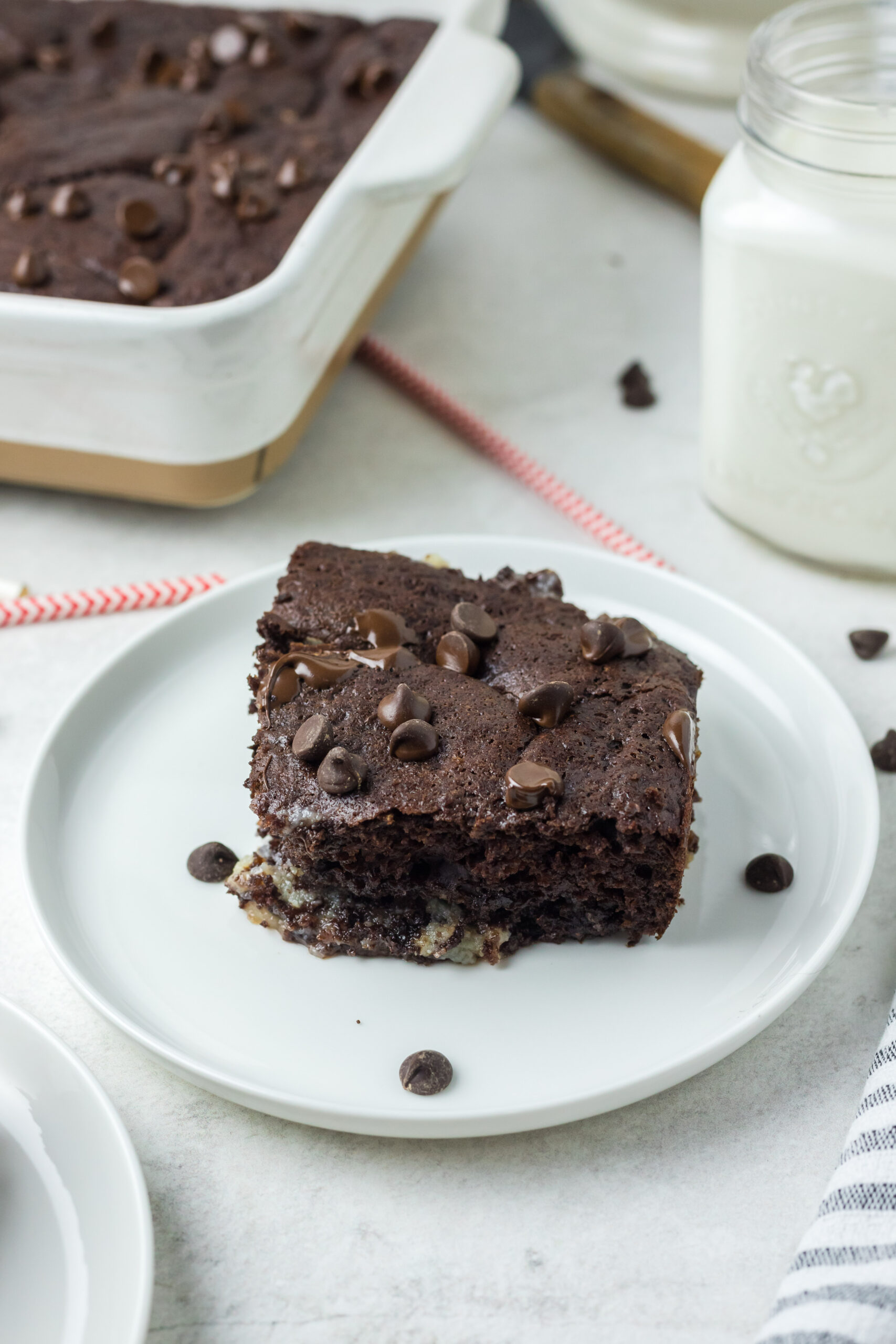 This is a pitcture of a piece of the chocolate earthquake cake with sprinkles of chocolate chips a glass of milk and the remainder of the cake.  The piece of cake is positioned in the front.