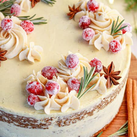 A frosted spice layer cake topped with eggnog buttercream swirls and sugared cranberries on a cake stand.