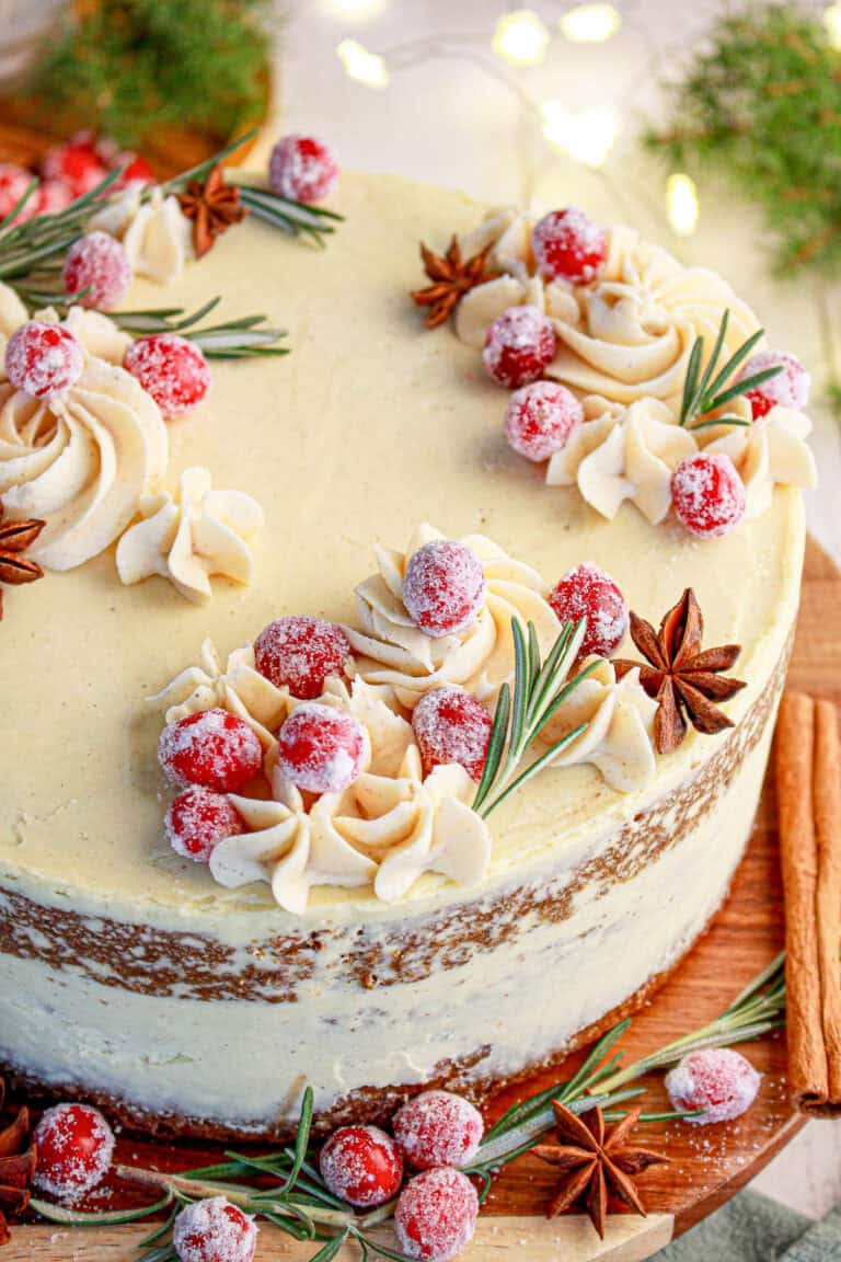 A frosted spice layer cake topped with eggnog buttercream swirls and sugared cranberries on a cake stand.