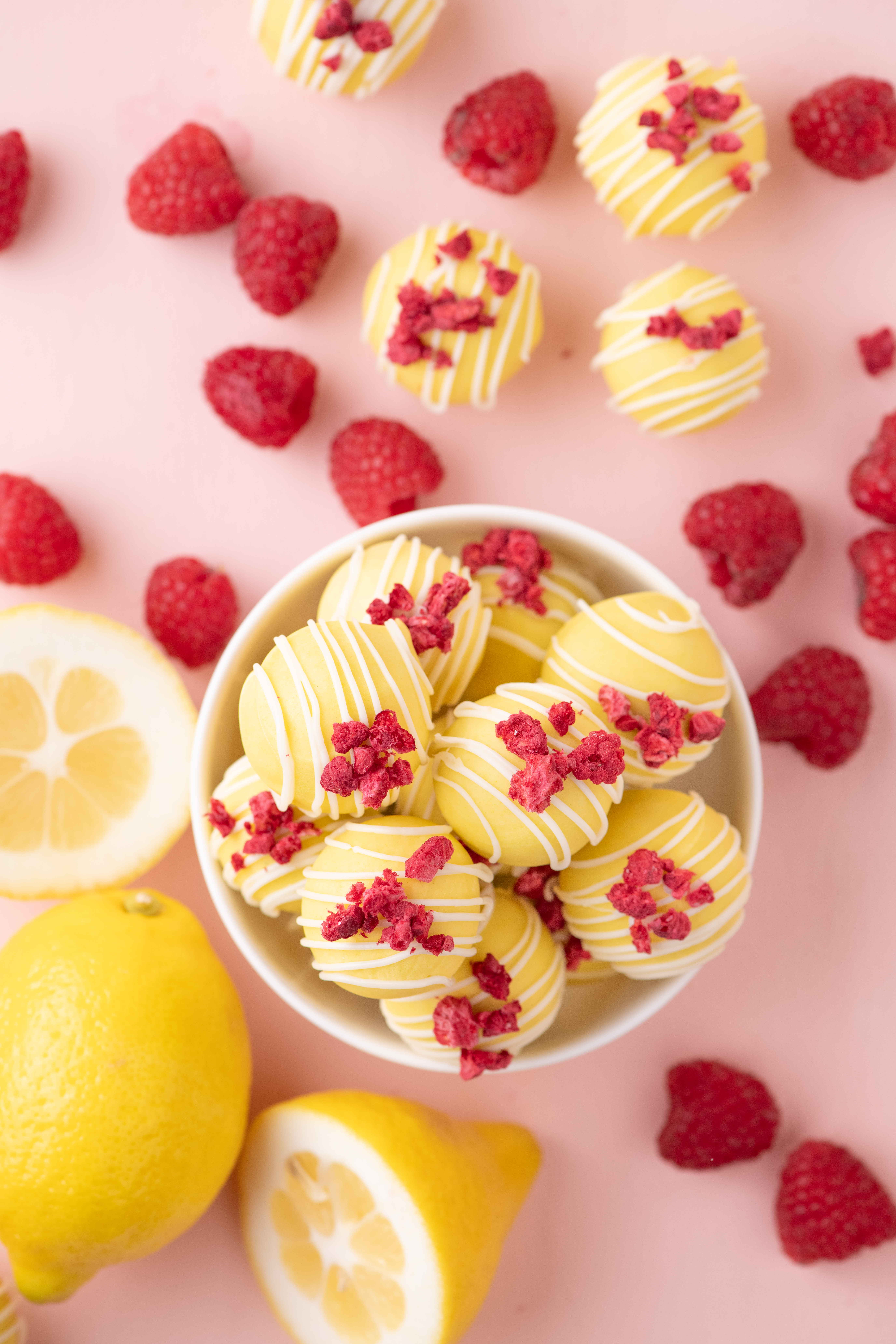 Bowl of lemon raspberry cake balls topped with white chocolate drizzle and raspberry crumble with fresh lemons and raspberries.