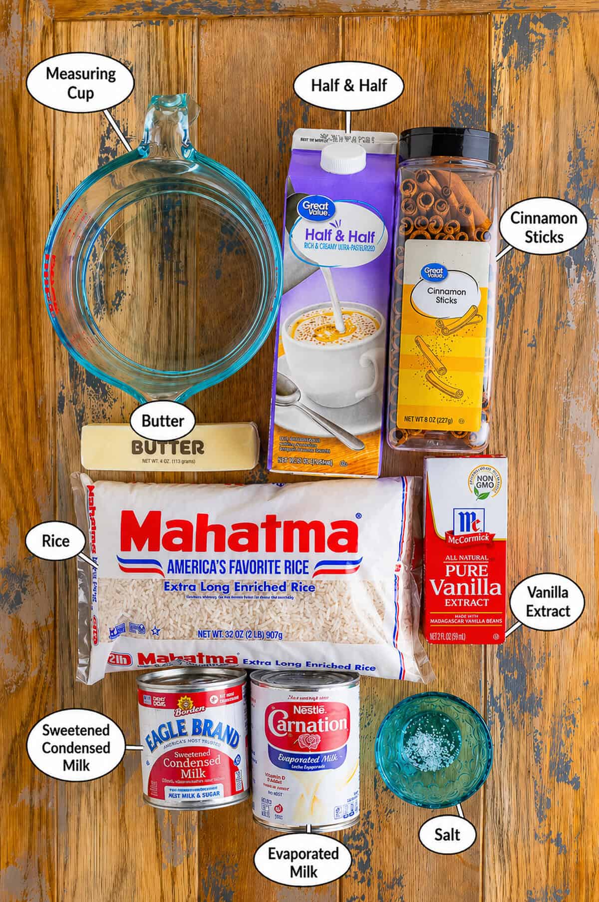 Mexican rice pudding ingredients arranged on rustic wood, including rice, milk, cinnamon, vanilla, butter, salt, and half and half cream.