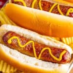 A pair of Air Fryer Cheddar Brats nestled on soft buns with mustard and grilled onions on a red checkered cloth.