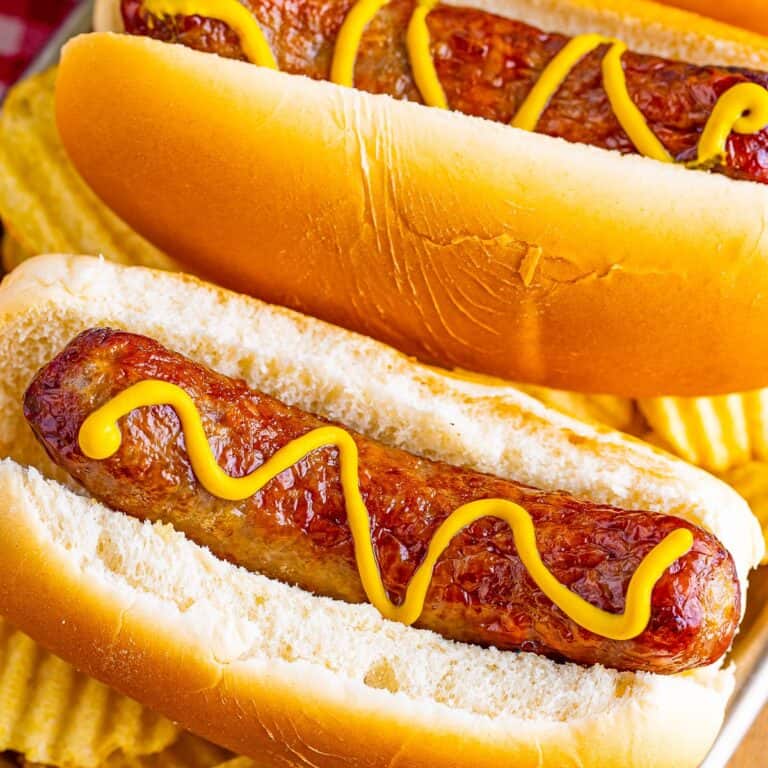 A pair of Air Fryer Cheddar Brats nestled on soft buns with mustard and grilled onions on a red checkered cloth.