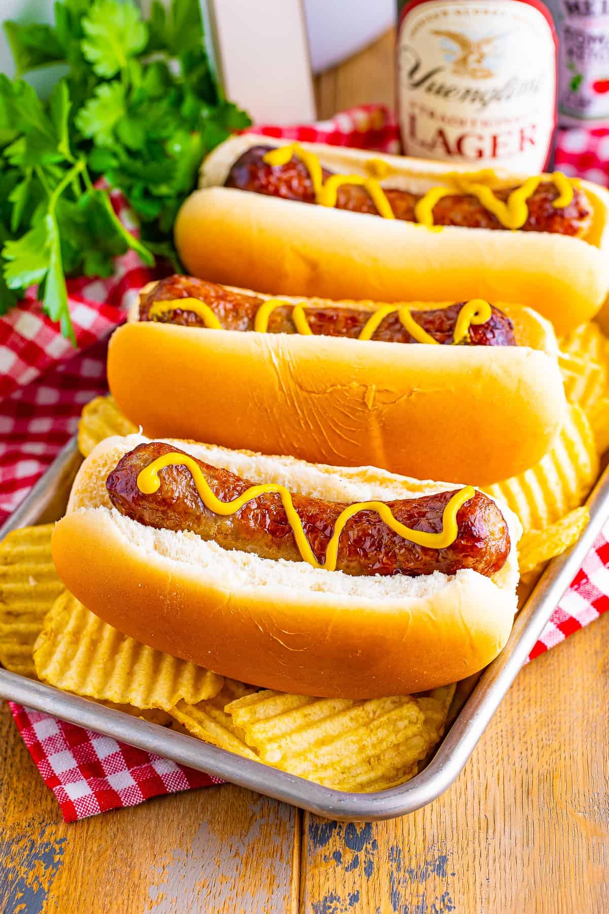 Two Johnsonville Brats in Air Fryer served on toasted buns with mustard, fresh onions, and herbs on a red checkered linen.