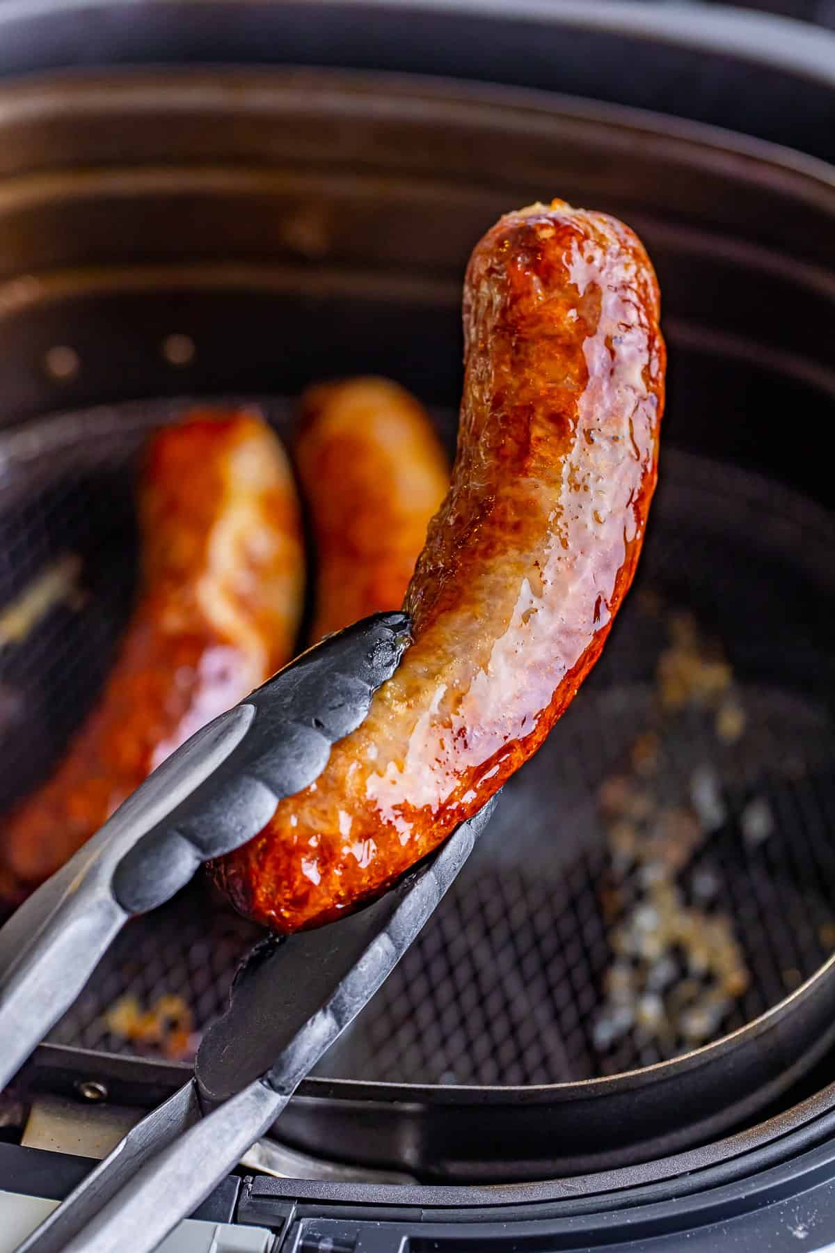 A pair of cooked Johnsonville Brats in Air Fryer lifted from the basket with kitchen tongs.