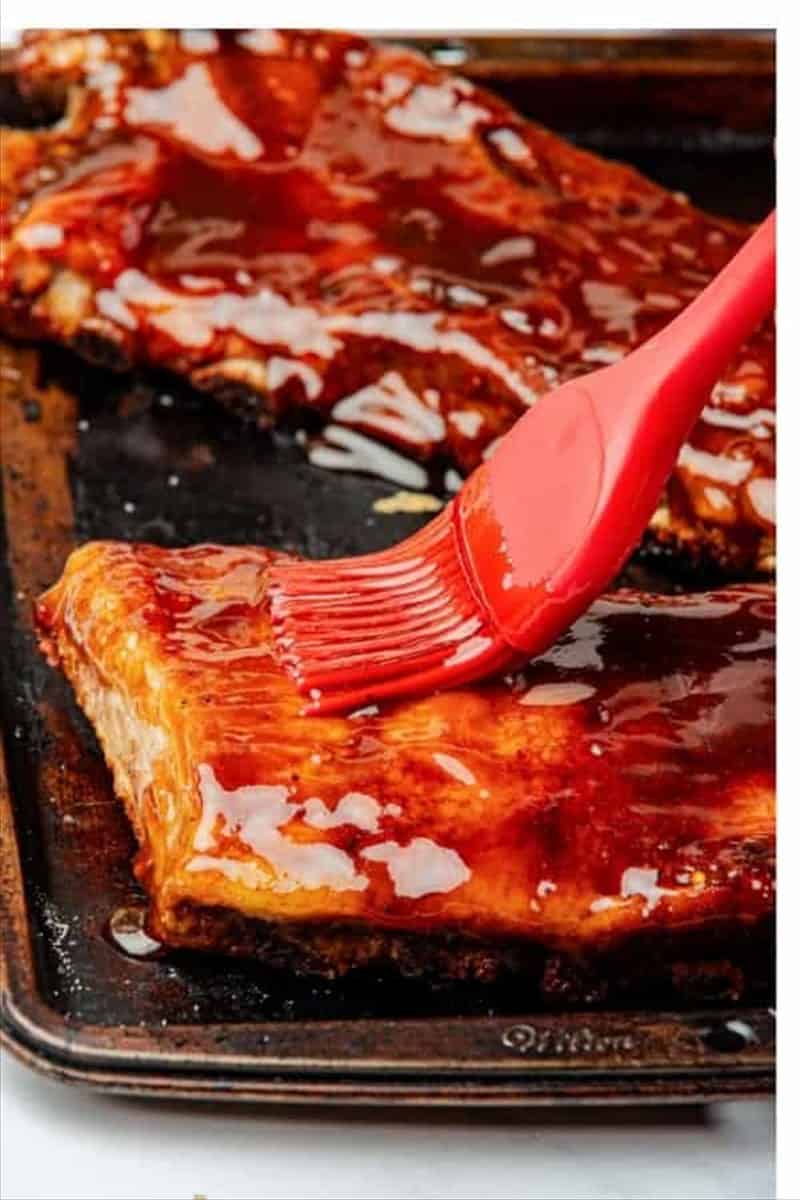 Red silicone pastry brush coating partially baked St Louis Ribs with BBQ honey glaze on a baking tray for Best Oven St Louis Ribs Recipe.