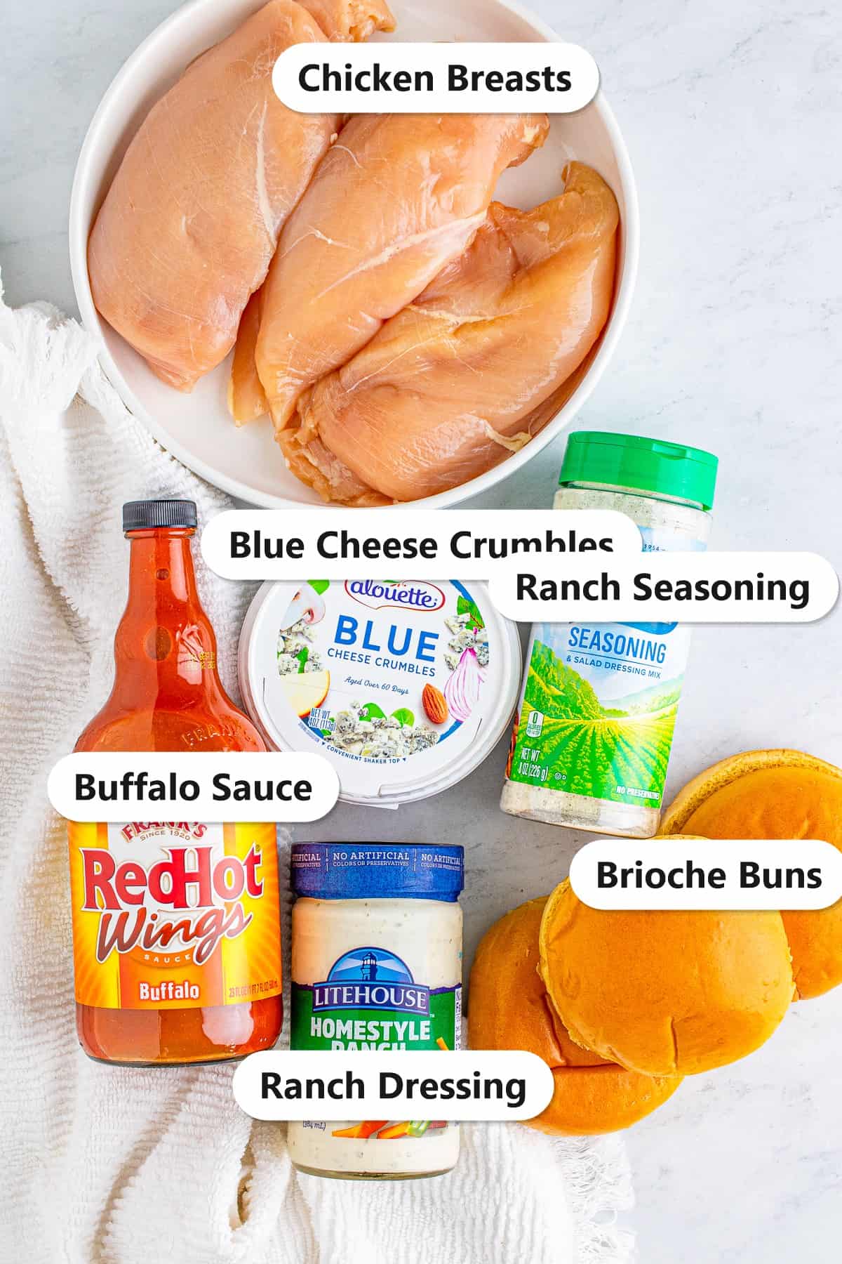 Labeled ingredients for slow cooker buffalo chicken sandwiches including chicken breasts, buffalo sauce, ranch seasoning, blue cheese, ranch dressing, and brioche buns.