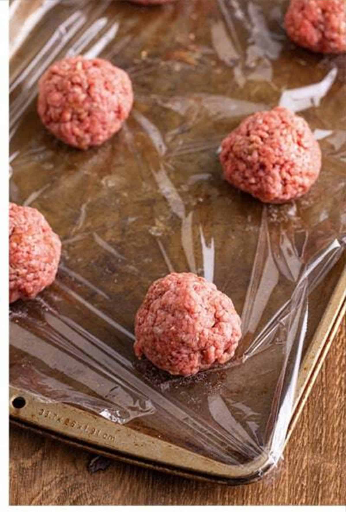 Six portioned balls of seasoned ground beef on plastic wrap for cheeseburger sliders.