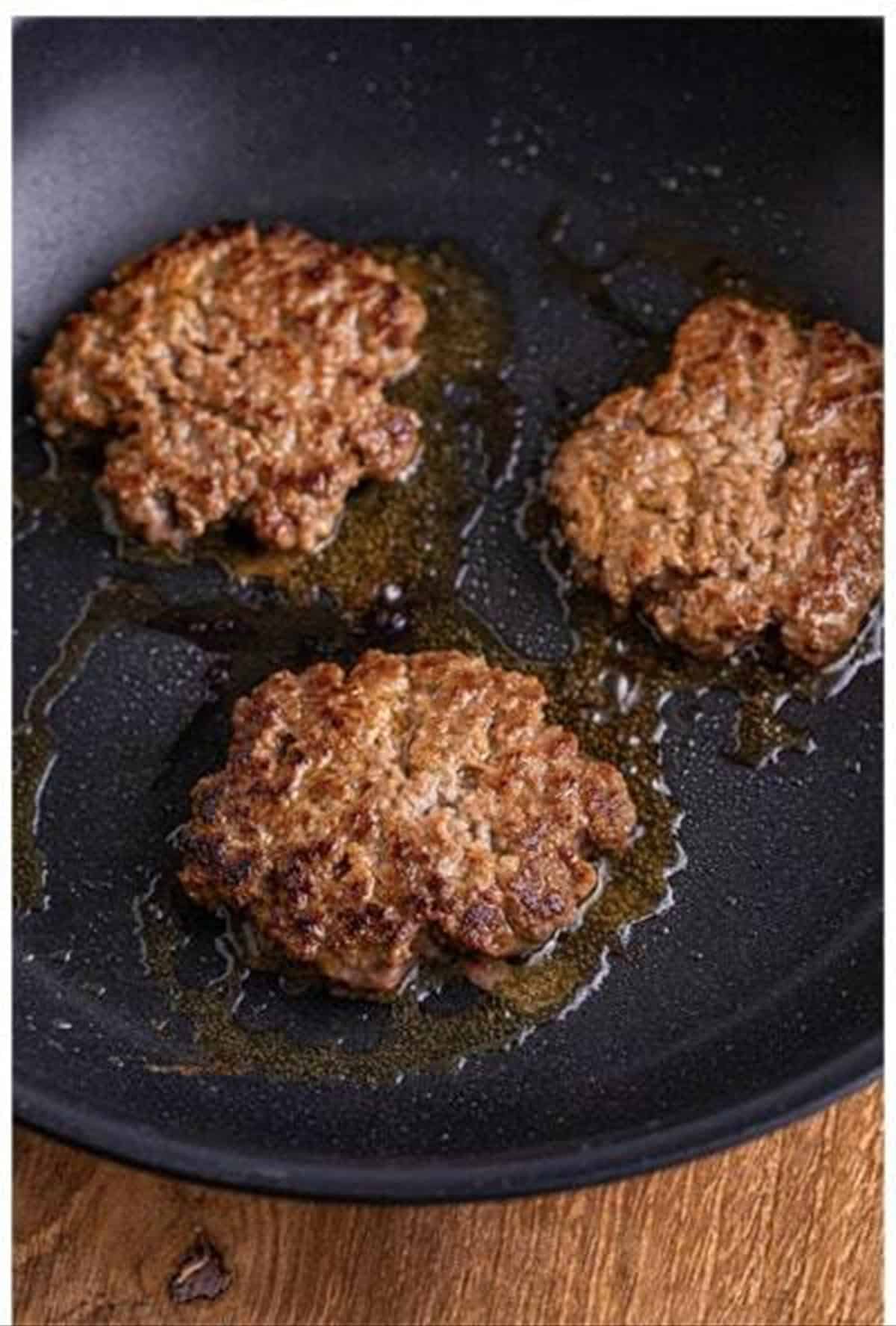 Cheeseburger slider patties cooking and forming a crust in a black skillet.