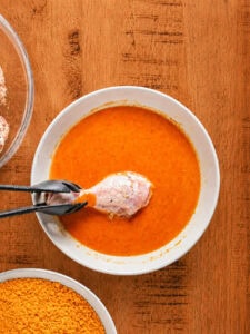 Chicken piece being dipped into the buffalo egg wash for cheesy fried chicken.