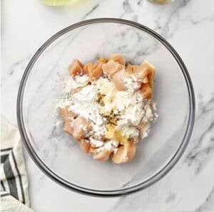 Raw diced chicken pieces in a glass mixing bowl with flour, cornstarch, and seasonings poured over top, ready to be tossed for coating before frying.