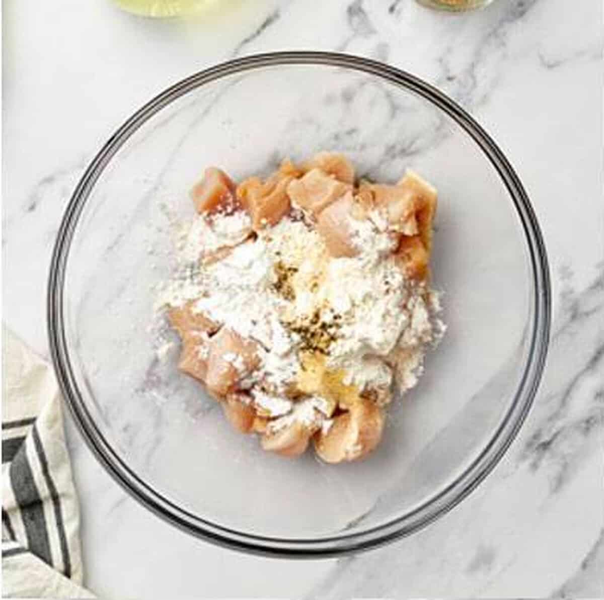 Raw diced chicken pieces in a glass mixing bowl with flour, cornstarch, and seasonings poured over top, ready to be tossed for coating before frying.
