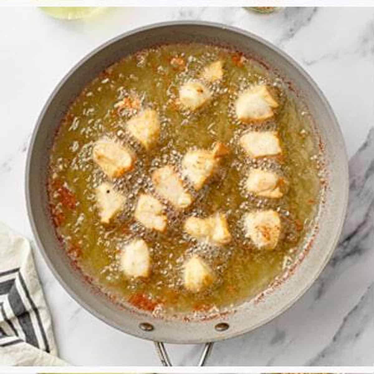 Battered Chinese Chicken bites frying in hot bubbling oil in a large silver skillet, cooking until golden and crispy on the outside, overhead view on marble.