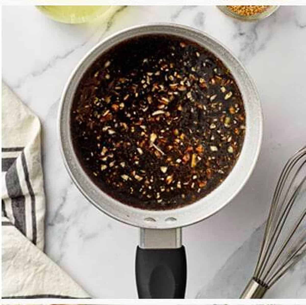 Dark brown sugar soy garlic glaze bubbling and simmering in a small silver saucepan with minced garlic, a metal whisk resting beside it on marble.