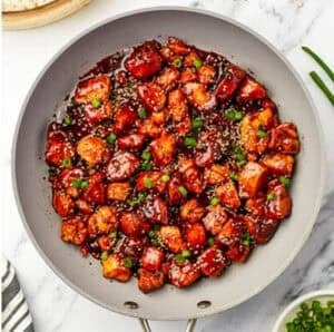 Crispy Chinese Chicken bites fully coated and glossy with dark brown sugar soy glaze, topped with sesame seeds and green onions in a large gray skillet.