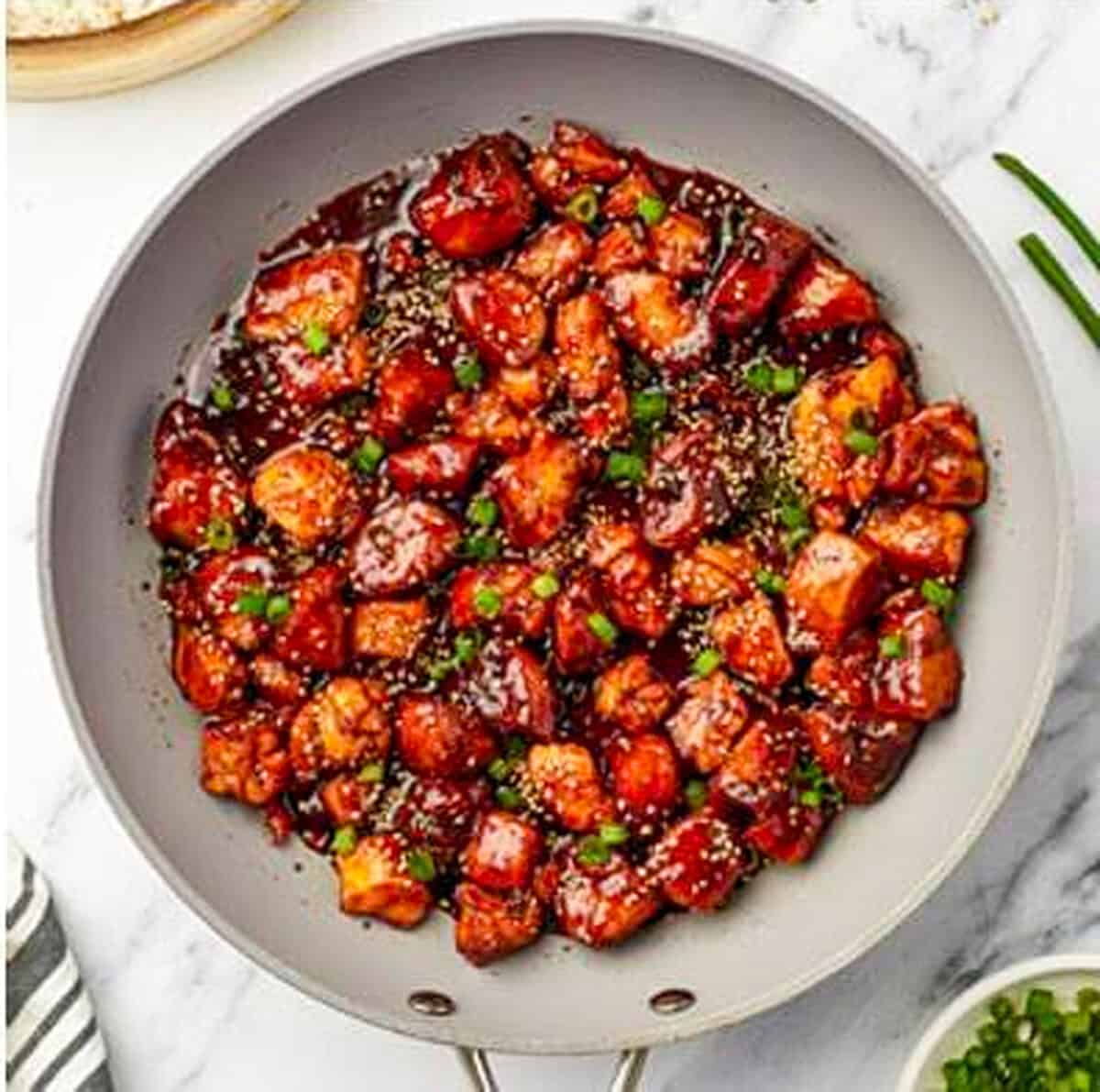 Crispy Chinese Chicken bites fully coated and glossy with dark brown sugar soy glaze, topped with sesame seeds and green onions in a large gray skillet.