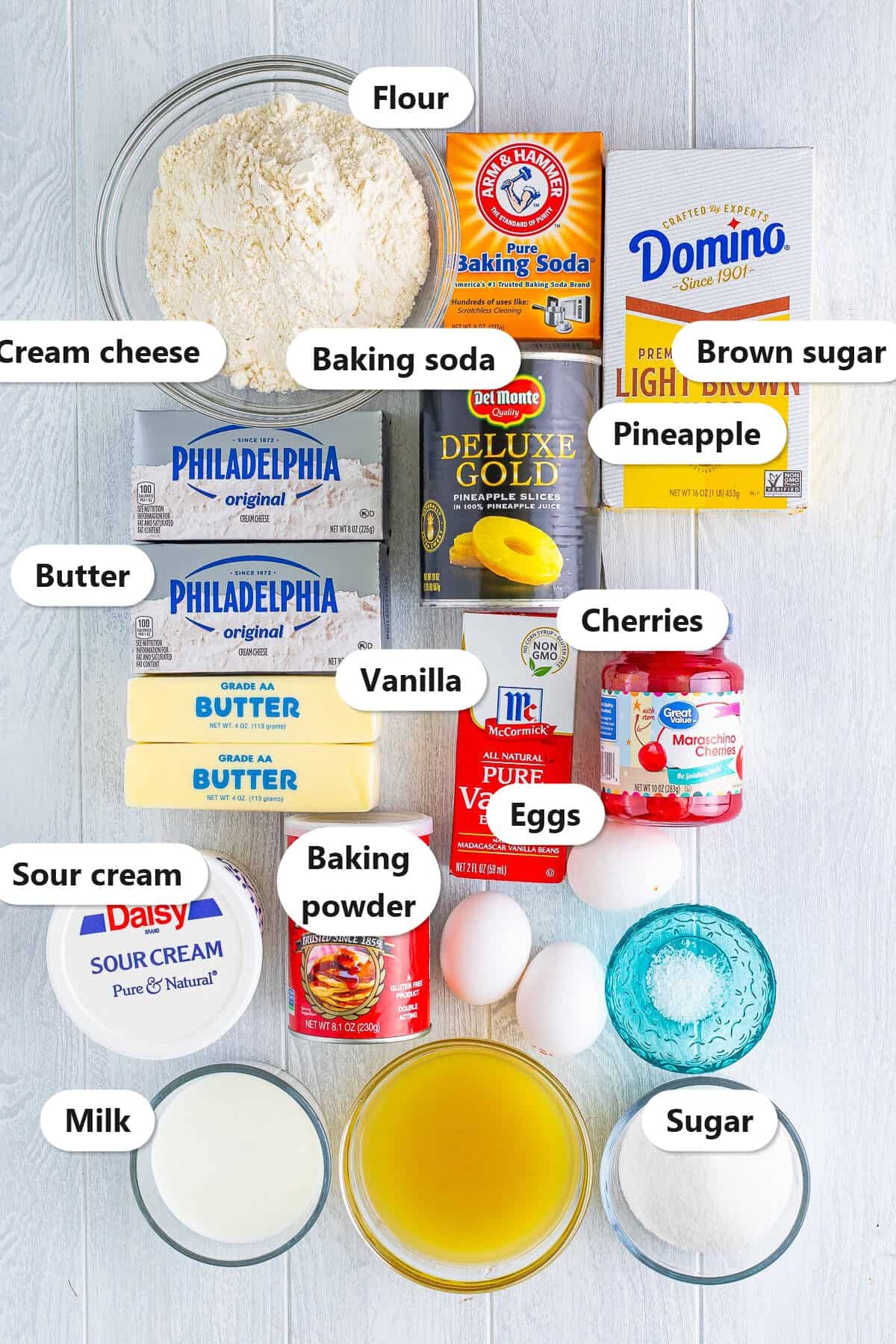 Labeled ingredient flat lay for Copycat Cheesecake Factory Pineapple Upside Down Cheesecake showing flour, baking soda, brown sugar, cream cheese, pineapple, butter, vanilla, cherries, sour cream, baking powder, eggs, milk, and sugar.