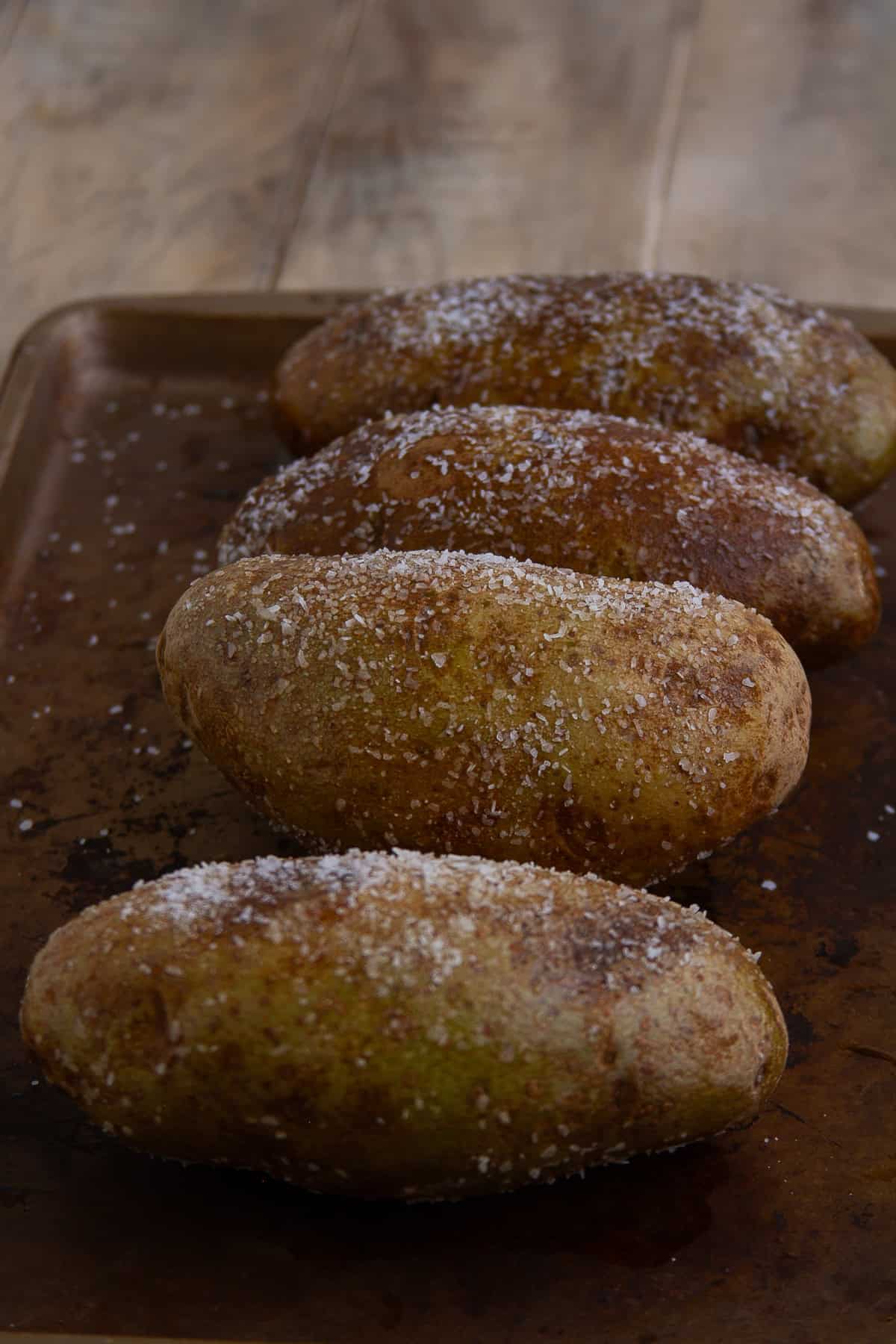 Four russet potatoes generously coated with olive oil and kosher salt on a baking sheet ready to bake into Crispy Baked Potato.