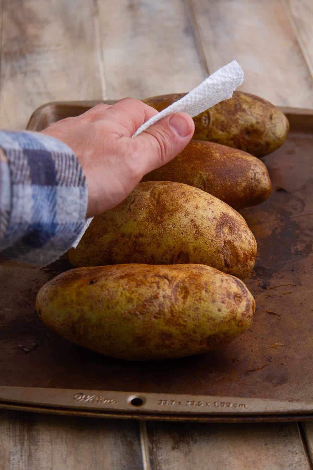 Hand patting freshly scrubbed russet potatoes dry with a paper towel on a baking sheet for Crispy Baked Potato.