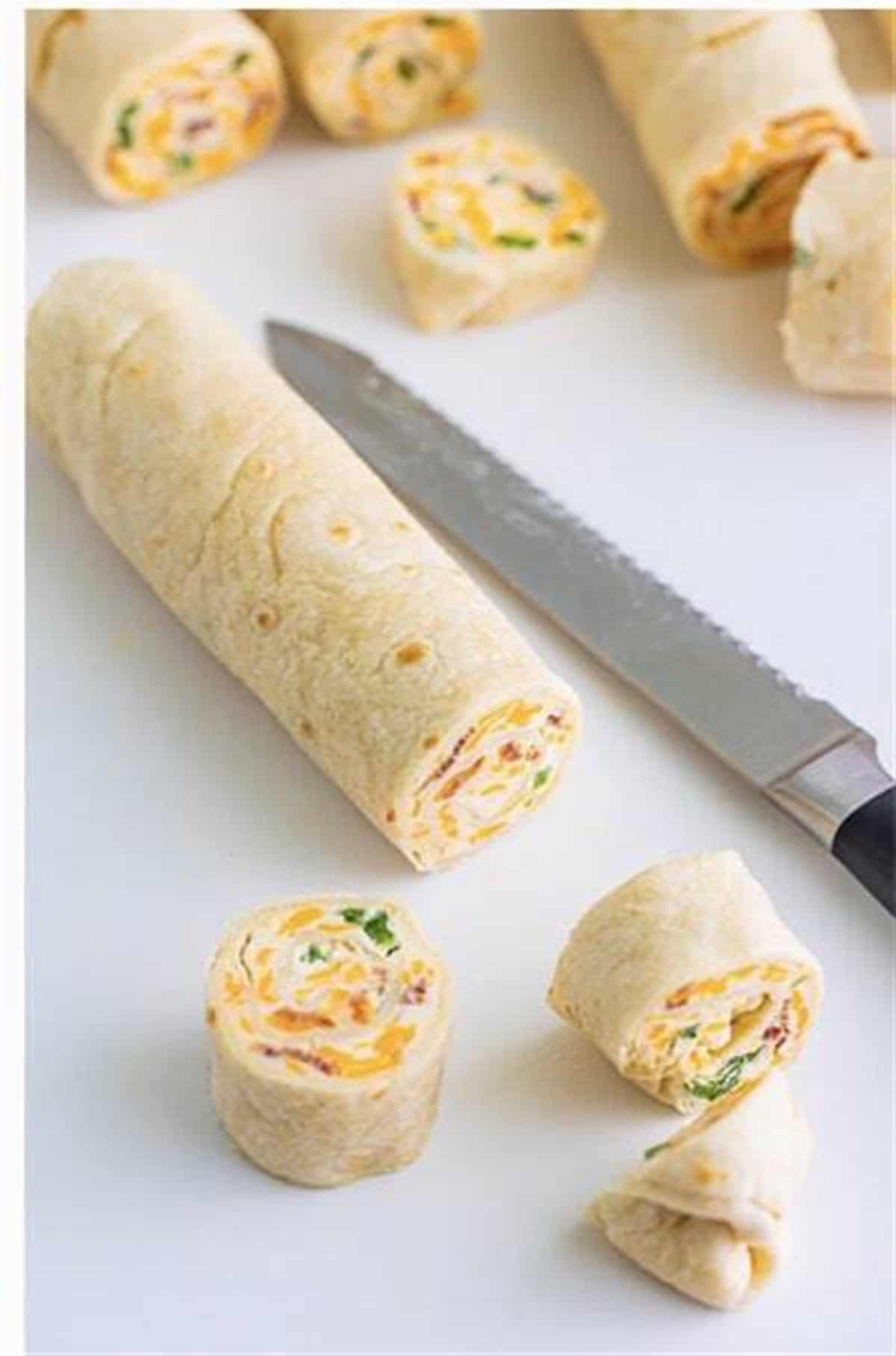 Rolled tortilla log being sliced into half-inch Easy Cream Cheese Pinwheels with a serrated knife.