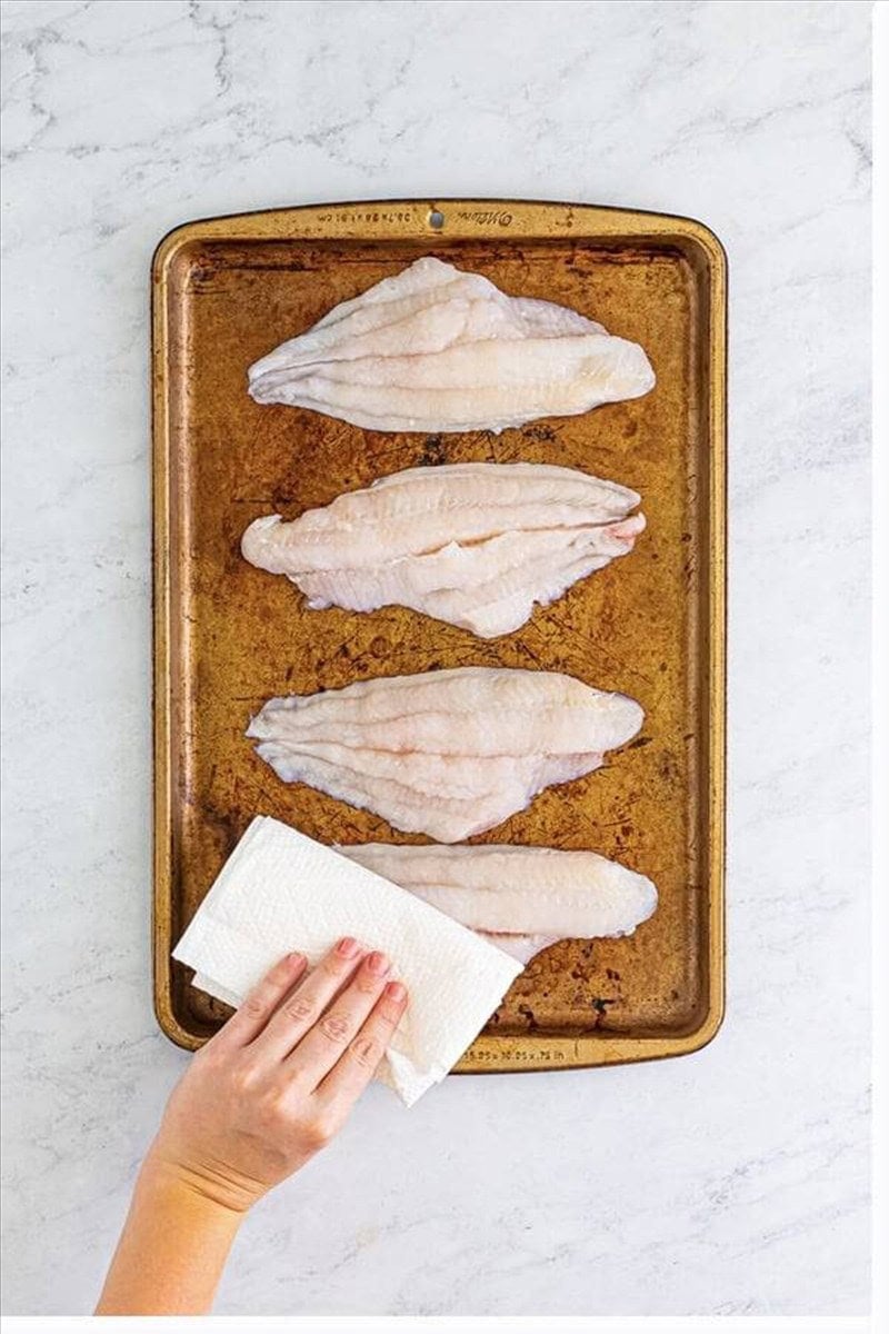 Hand patting raw catfish filets dry with paper towels on a sheet tray before frying.